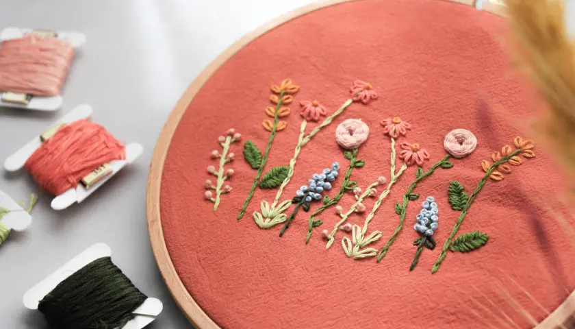 Zarte Stickereien auf Textilien zeigen eine Reihe von Blumen und Blättern in einem Stickrahmen. Fadenrollen liegen daneben.