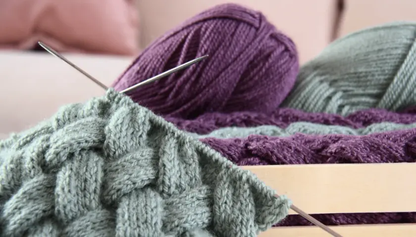 Stricken mit dicker Wolle: Ein Strickprojekt mit Zopfmuster in Grau und Lila, Nadeln stecken im Gestrick.