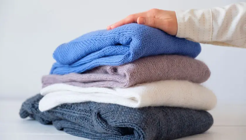 Ein Stapel gestrickter Pullover in verschiedenen Farben, von dunkelblau bis weiß. Eine Hand streicht sanft über den obersten Pullover, als ob er per Hand gewaschen wurde.