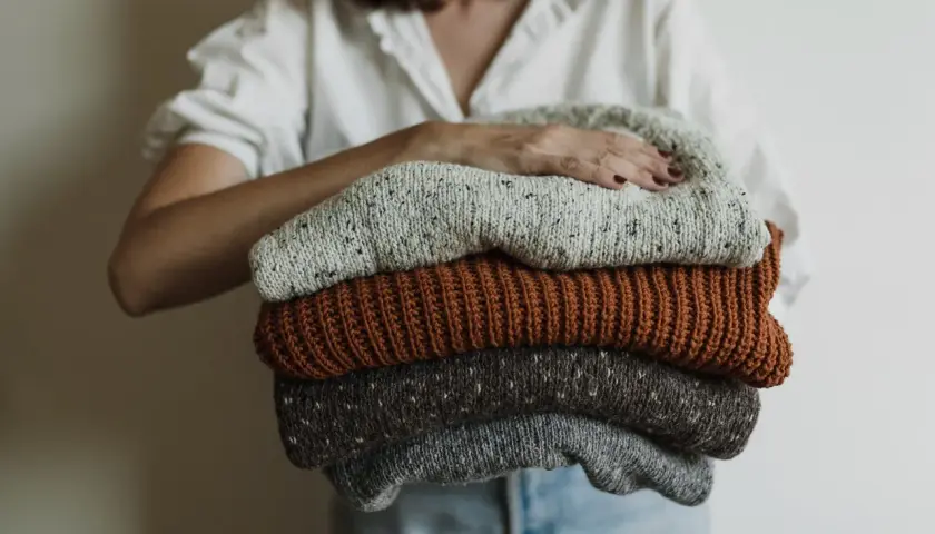 Eine Person hält einen Stapel gestrickter Pullover. Die Pullover sind in Grau-, Braun- und Beigetönen gehalten. Sie sind bereit, um **Wolle bei 40 Grad waschen** zu können.
