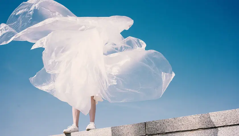 Eine Person steht auf einer Mauer, umgeben von wehendem, weißem Material, das an Plastik erinnert. Polyester schädlich für die Umwelt.