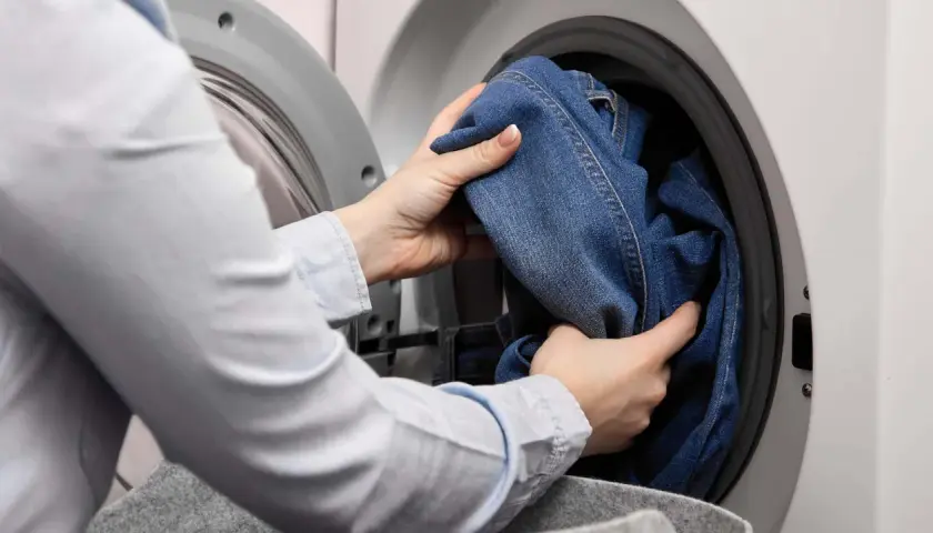Hände legen Jeans in die Waschmaschine. So kannst du deine Jeans richtig waschen und sie bleiben lange schön.