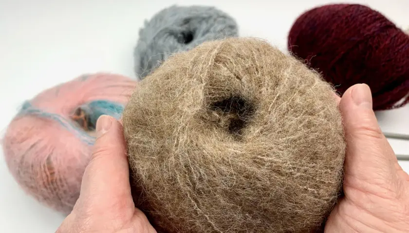 Hände halten ein Knäuel beiger Wolle, bereit, einen Schal zu stricken. Weitere Wollknäuel in Rosa, Grau und Bordeauxrot liegen im Hintergrund.