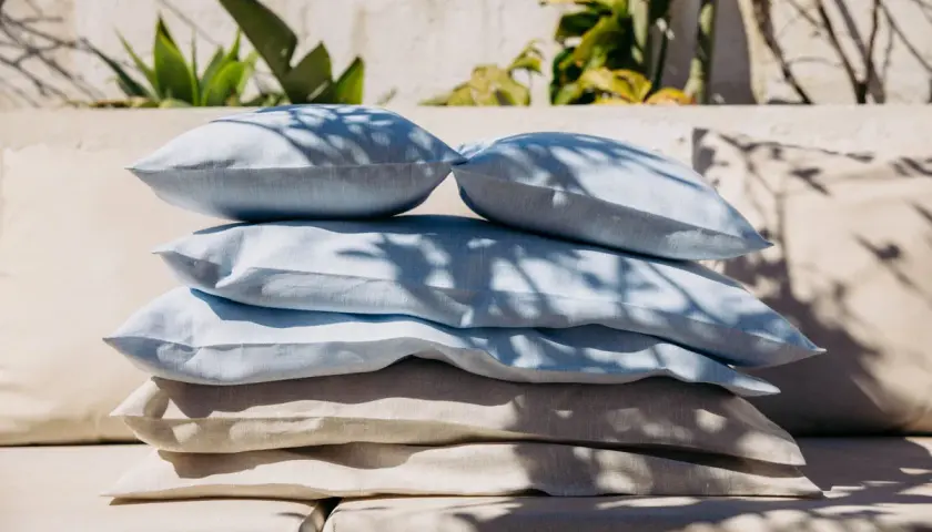 Stapel Kissen in Blau und Beige. Der feine Unterschied zwischen Leinen und Baumwolle ist spürbar. Schatten von Blättern fallen darauf.