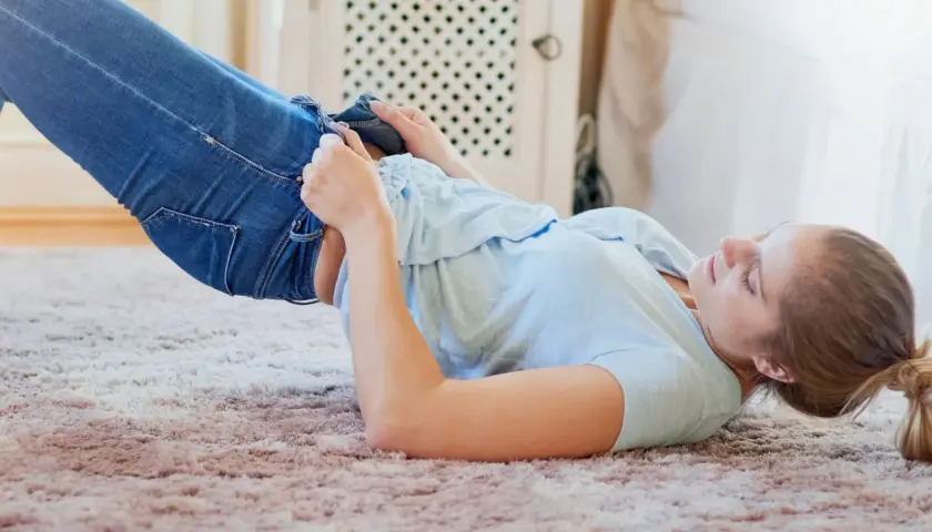 Frau kämpft mit ihrer Jeans, die eingelaufen ist. Was tun? Sie liegt auf einem Teppich und versucht, die Hose anzuziehen.