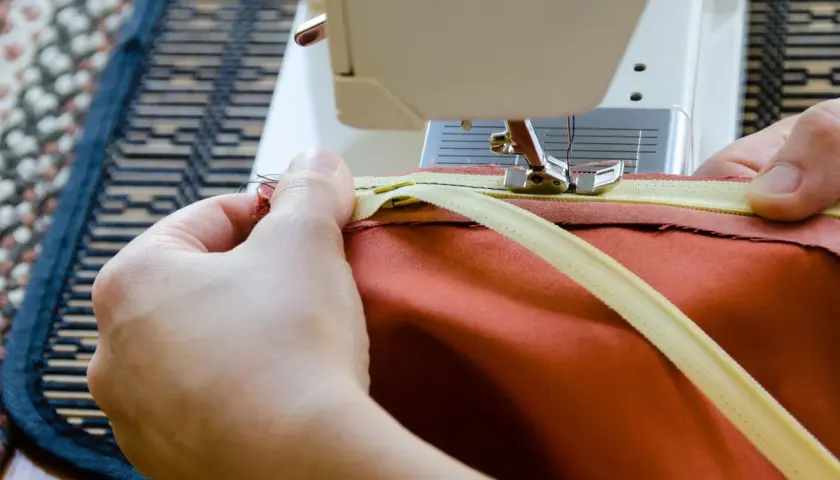 Hände nähen einen Reißverschluss mit einer Nähmaschine an ein orangefarbenes Kleidungsstück. Reißverschluss reparieren leicht gemacht.