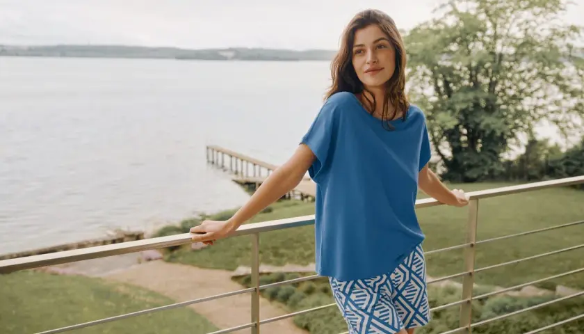 Frau auf Balkon mit Blick aufs Wasser, trägt blaues Oberteil und gemusterte Shorts aus Modal Stoff. Waschen ist einfach.