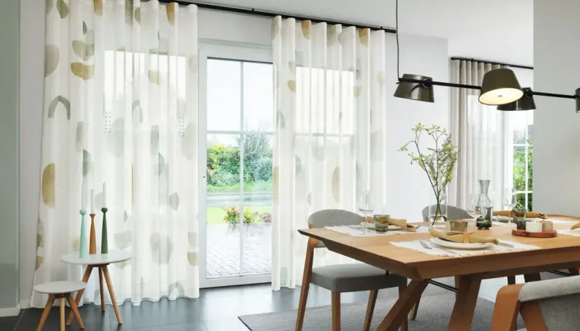 Ein Esszimmer mit Holztisch und Stühlen. Ein Vorhang nur auf einer Fensterseite mit abstrakten Mustern.