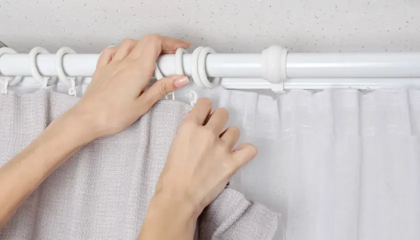 Hände befestigen zwei Gardinen auf einer Stange. Eine Gardine ist grau, die andere weiß und transparent.