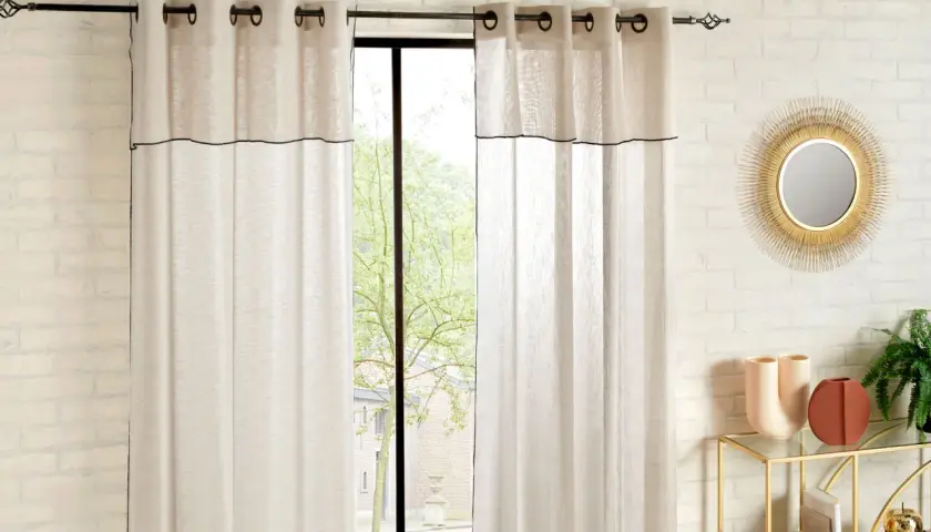 Helle Vorhänge mit schwarzer Umrandung sind an einer Wand befestigt. Ein Fenster mit Blick ins Grüne ist teilweise verdeckt.