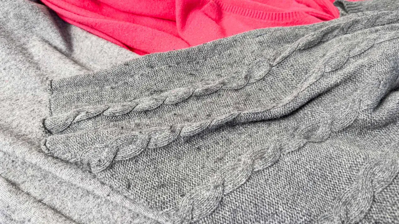 Grauer Strickpullover mit Zopfmuster und ein pinker Stoff. Kleine Knötchen zeigen, wie man Pilling vermeiden kann.