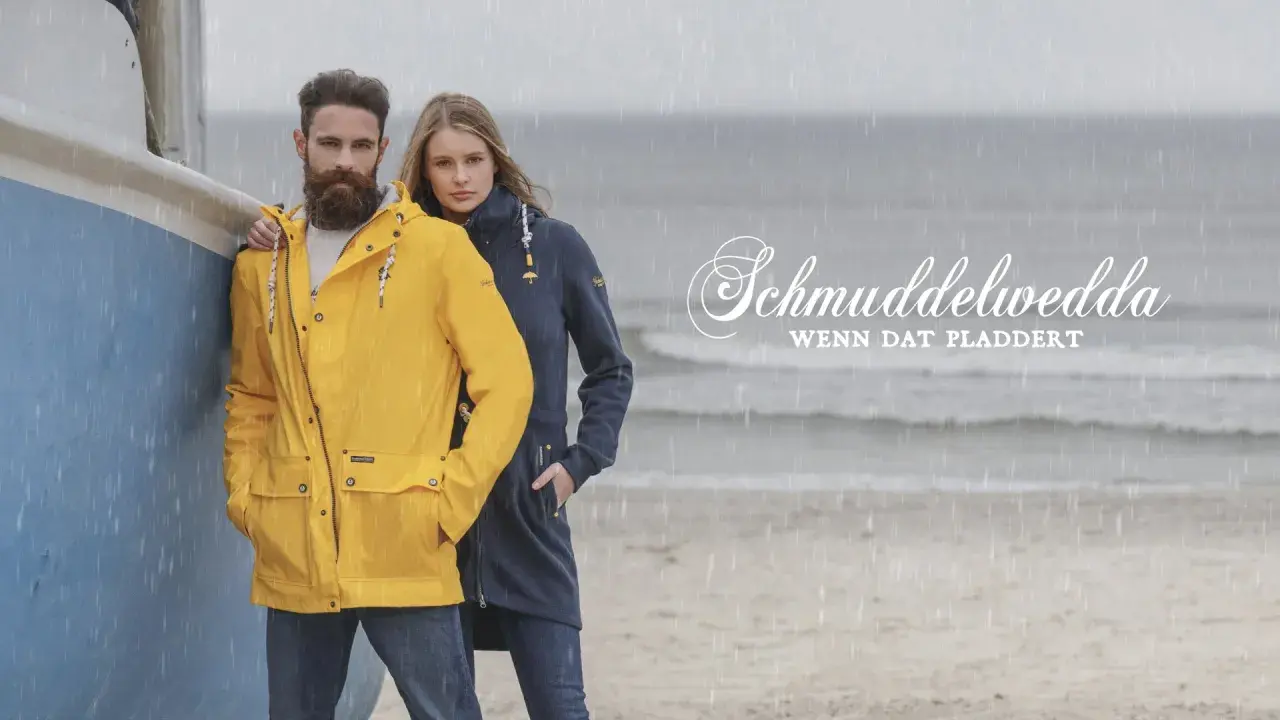 Mann und Frau am Strand im Regen, er trägt eine gelbe Schmuddelwedda Jacke, sie eine blaue. Perfekt für jedes Wetter!