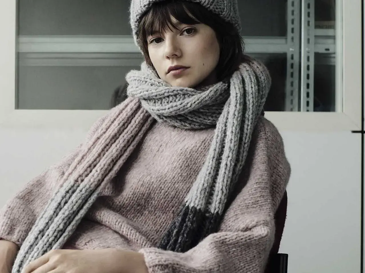 Junges Mädchen mit einem gestrickten Schal aus Wolle und passender Mütze. Der dicke, graue Schal ist um den Hals geschlungen und reicht bis zum rosafarbenen Pullover.
