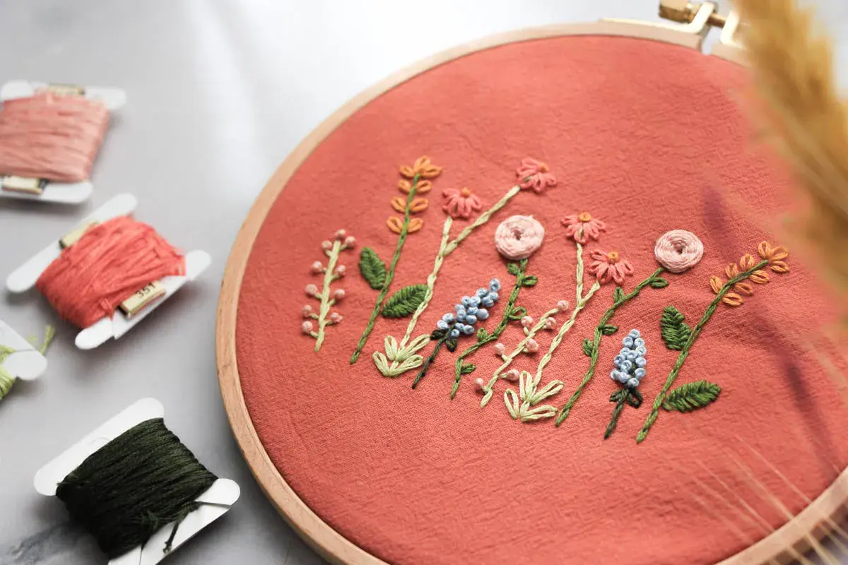 Zarte Stickereien auf Textilien zeigen eine Reihe von Blumen und Blättern in einem Stickrahmen. Fadenrollen liegen daneben.