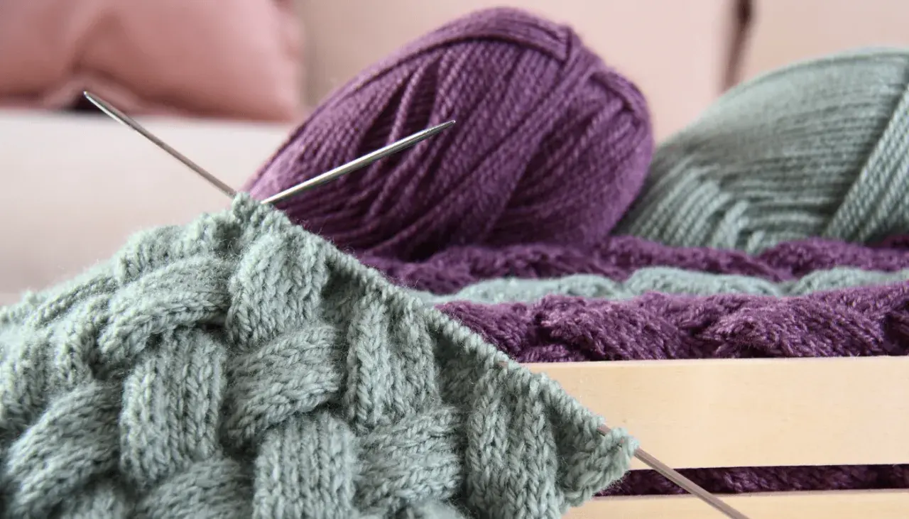 Stricken mit dicker Wolle: Ein Strickprojekt mit Zopfmuster in Grau und Lila, Nadeln stecken im Gestrick.