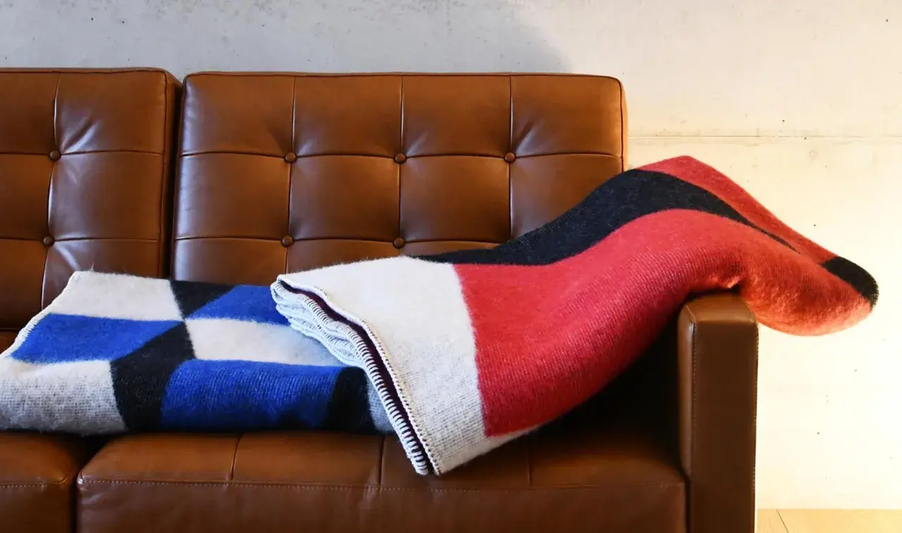Zwei Decken auf braunem Ledersofa: eine mit geometrischem Muster in Blau, Schwarz und Weiß, die andere in Rot, Schwarz und Weiß. Der Unterschied zwischen Wolle und Baumwolle ist spürbar.
