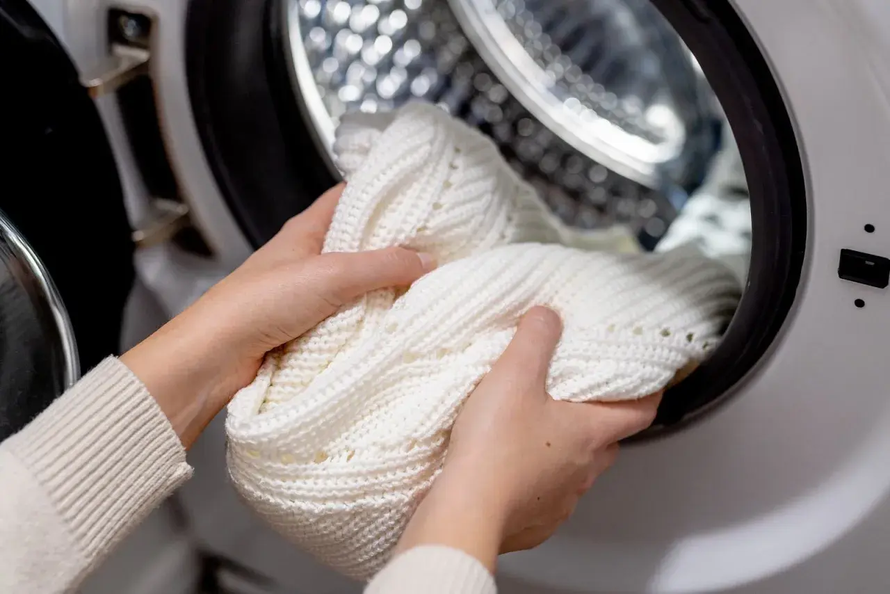 Hände legen einen weißen Strickpullover in die Waschmaschine, um eingelaufene Kleidung zu retten.