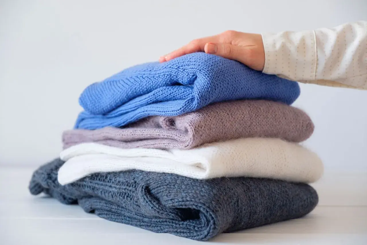 Ein Stapel gestrickter Pullover in verschiedenen Farben, von dunkelblau bis weiß. Eine Hand streicht sanft über den obersten Pullover, als ob er per Hand gewaschen wurde.