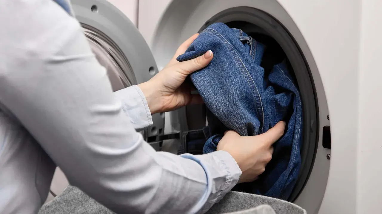 Hände legen Jeans in die Waschmaschine. So kannst du deine Jeans richtig waschen und sie bleiben lange schön.