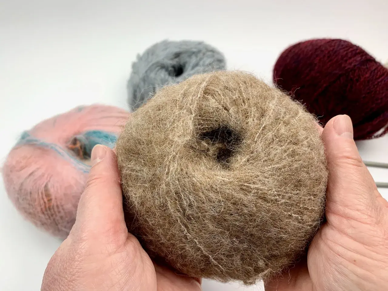 Hände halten ein Knäuel beiger Wolle, bereit, einen Schal zu stricken. Weitere Wollknäuel in Rosa, Grau und Bordeauxrot liegen im Hintergrund.