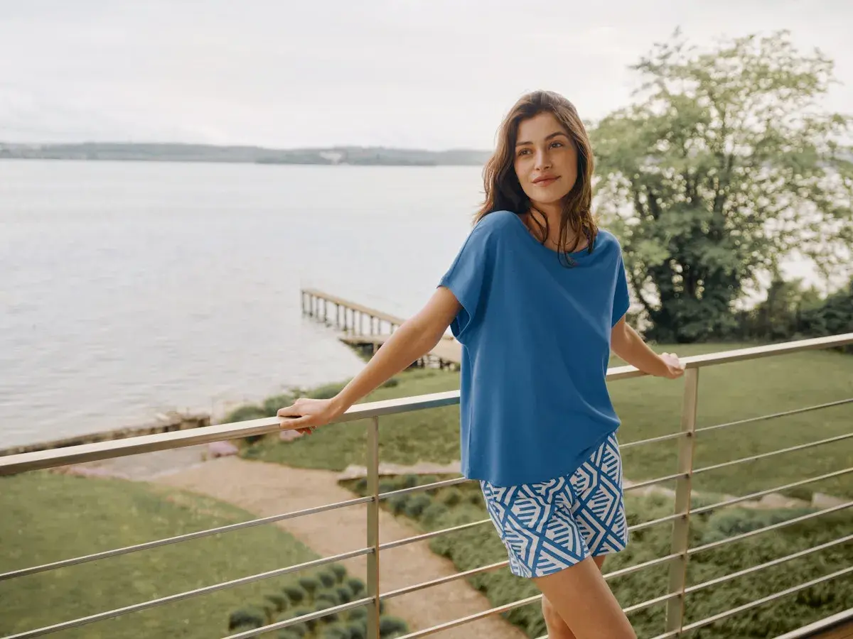 Frau auf Balkon mit Blick aufs Wasser, trägt blaues Oberteil und gemusterte Shorts aus Modal Stoff. Waschen ist einfach.