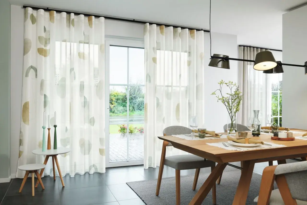 Ein Esszimmer mit Holztisch und Stühlen. Ein Vorhang nur auf einer Fensterseite mit abstrakten Mustern.