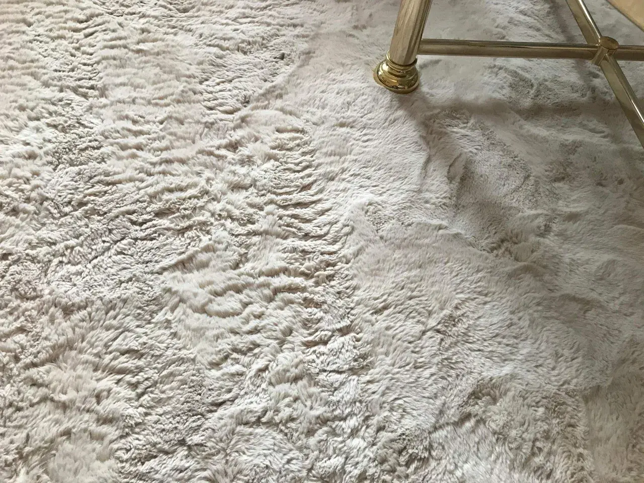 Ein heller, flauschiger Kunstfell Teppich ist leicht verfilzt. Ein goldener Möbelfuß steht darauf.