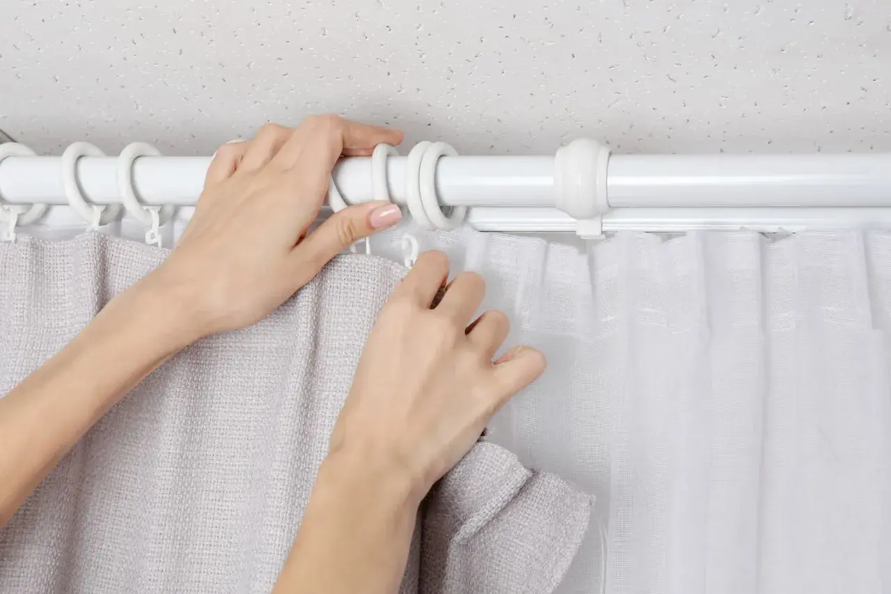 Hände befestigen zwei Gardinen auf einer Stange. Eine Gardine ist grau, die andere weiß und transparent.
