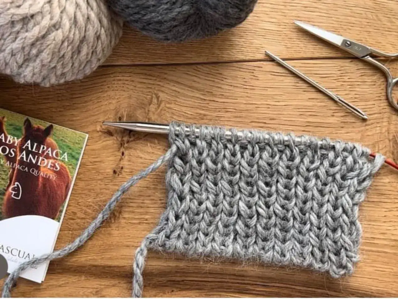 Strickprojekt mit grobem Garn, Nadeln, Schere und Alpaka-Garnknäueln auf Holzhintergrund.