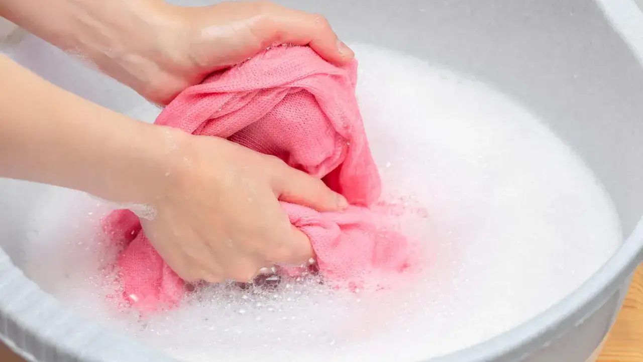 Hände waschen einen rosafarbenen Pullover in schaumigem Wasser. Ein sanfter Wollprogramm-Waschgang ist hier die beste Wahl.