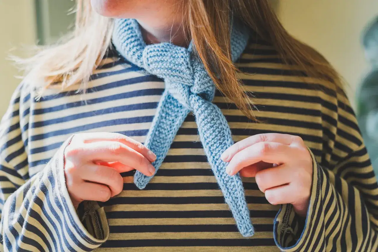 Ein hellblau gestrickter Schal mit einem einfachen Muster, um den Hals einer Person mit gestreiftem Oberteil geknotet.
