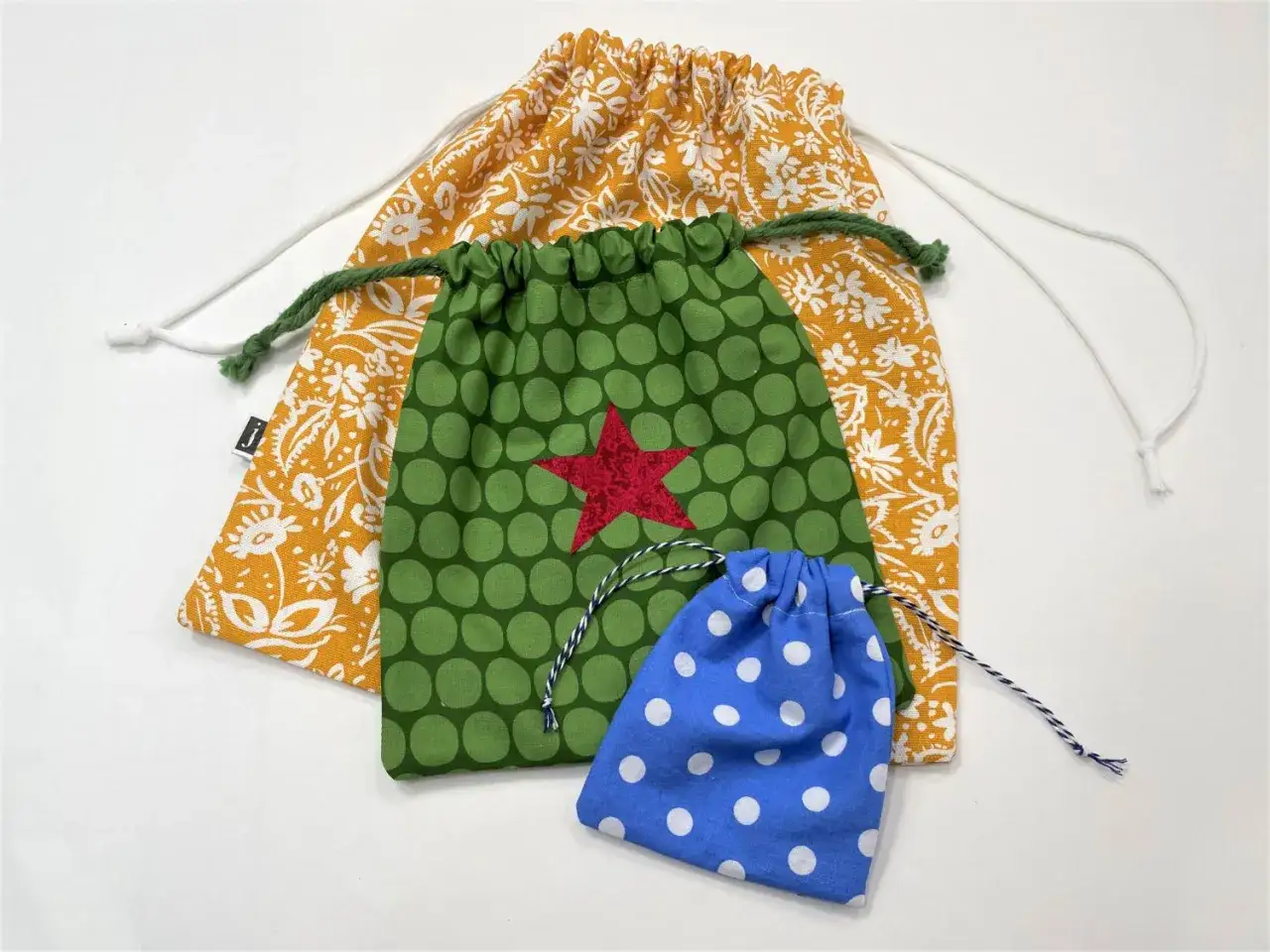 Drei bunte Beutel mit Kordelzug, inspiriert von einem Beutel Schnittmuster: gelb-wei&szlig; gemustert, gr&uuml;n mit roten Stern und blau mit wei&szlig;en Punkten.