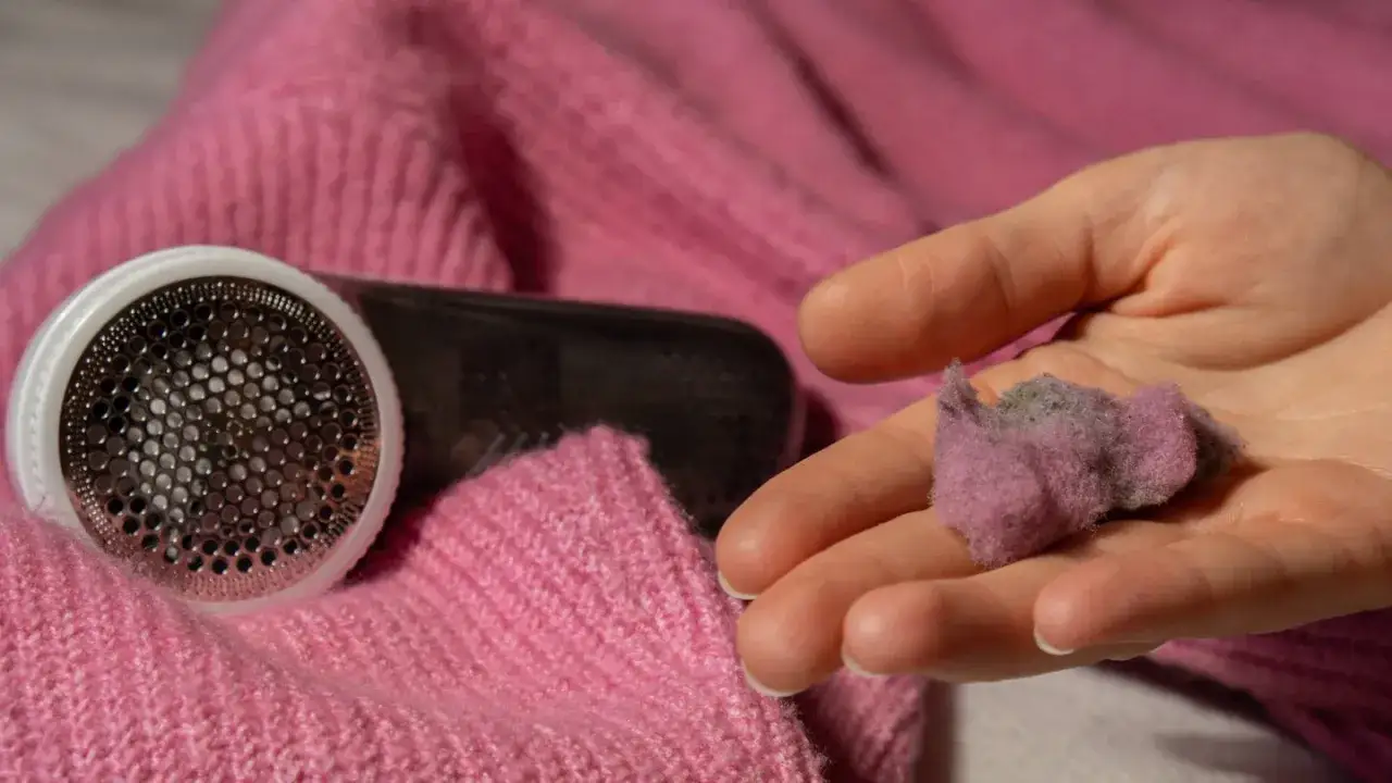 Ein Fusselrasierer liegt auf einem rosa Wollpullover. Eine Hand hält eine kleine Ansammlung von Fusseln, die beim wollpullover waschen entstanden sind.