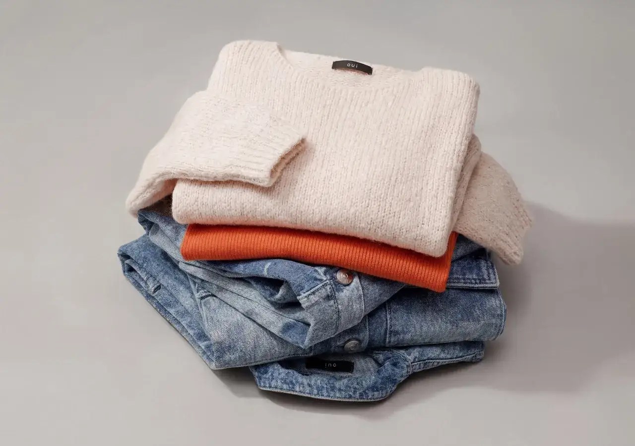 Ein Stapel Kleidung: cremefarbener wollpullover, orangefarbener Pullover und Jeansjacke. Perfekt für das wollpullover waschen.