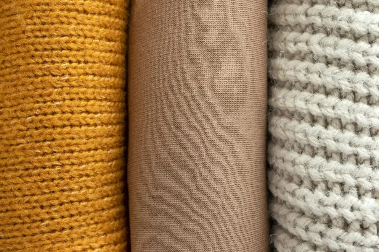 Drei Stoffrollen: gelb gestrickt, beige glatt und hellgrau grob gestrickt. Das beige Material k&ouml;nnte aus Polyester-Plastik sein.