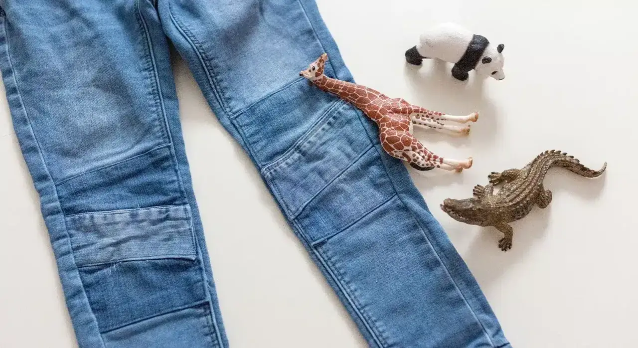 Jeans mit Flicken am Knie. Ein Spielzeug-Giraffe liegt auf dem Hosenbein, daneben ein Krokodil und ein Panda.