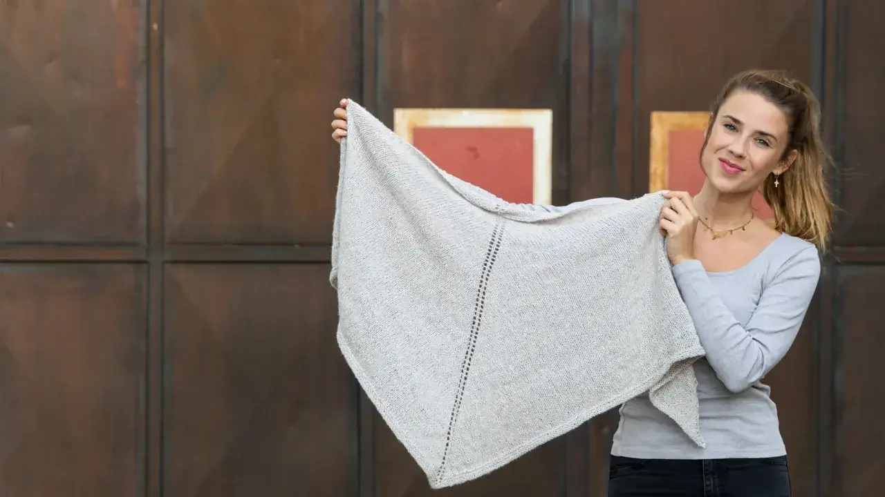 Junge Frau hält ein hellgraues, gestricktes Dreieckstuch hoch. Das Tuch hat ein schönes Muster und wurde mit viel Liebe zum Detail gestrickt.