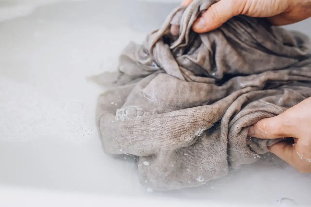 Hände waschen ein graubraunes Tuch in schaumigem Wasser. Sanfte Pflege für empfindliche Stoffe.