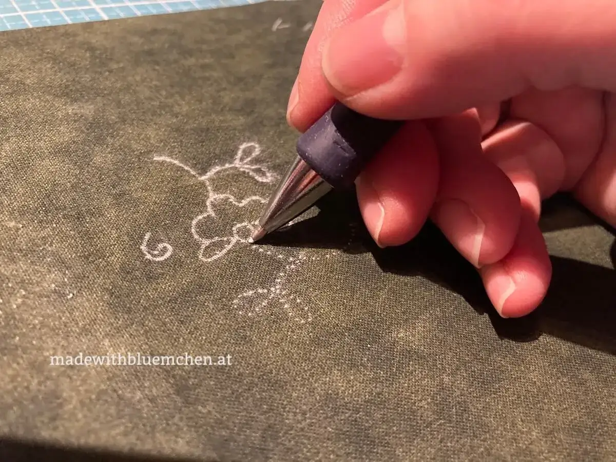 Hand überträgt ein zartes Blumenmuster auf Stoff. Das Stickmuster wird mit einem Stift auf den Stoff gezeichnet, bereit für die Stickerei.