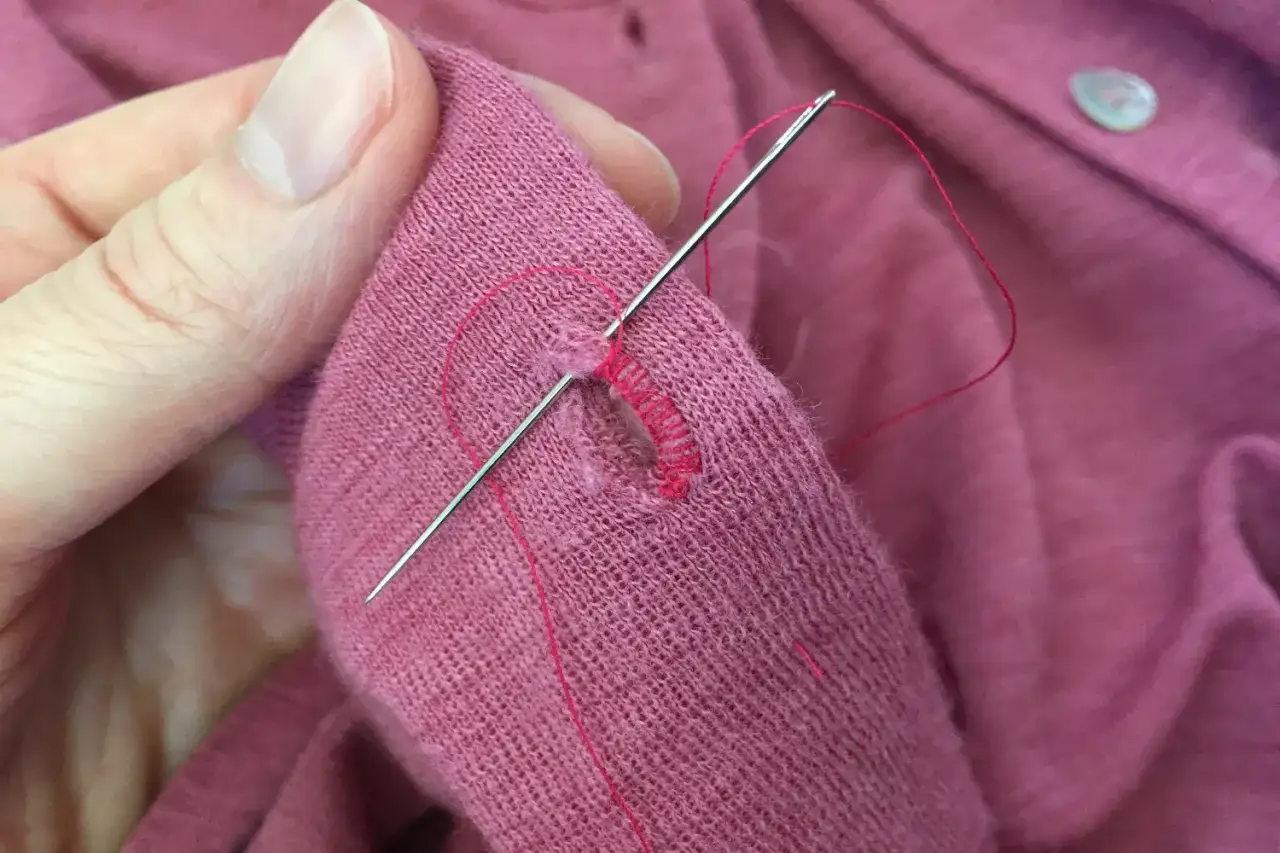 Eine Hand flickt mit rotem Garn ein Loch in einem rosafarbenen Kleidungsstück.